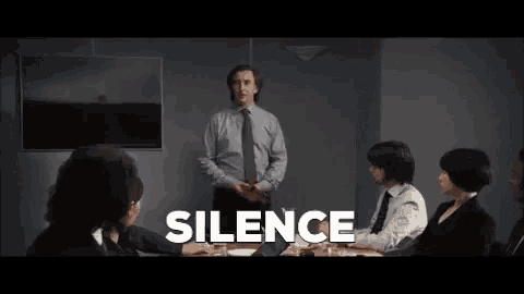 a man in a suit and tie is standing in front of a group of people sitting around a table and talking about silence .