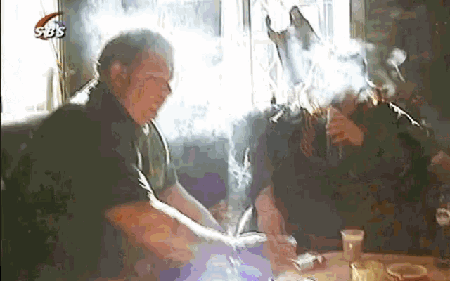 two men are sitting at a table with smoke coming out of their faces and a sbs logo in the corner