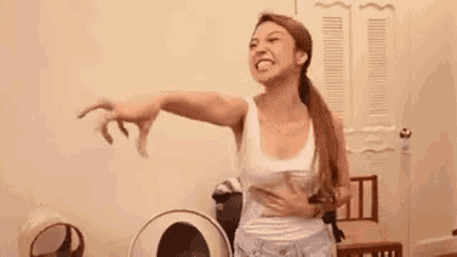 a woman in a white tank top is standing in a room with her arms outstretched and making a funny face .