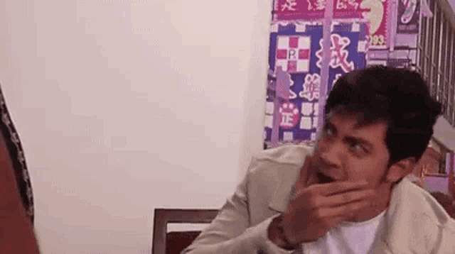 a man is covering his mouth with his hand while sitting in front of a wall with chinese writing on it .