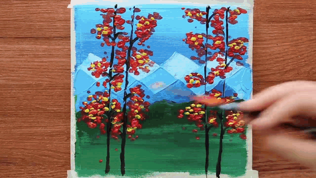a person is painting a picture of trees with mountains in the background .