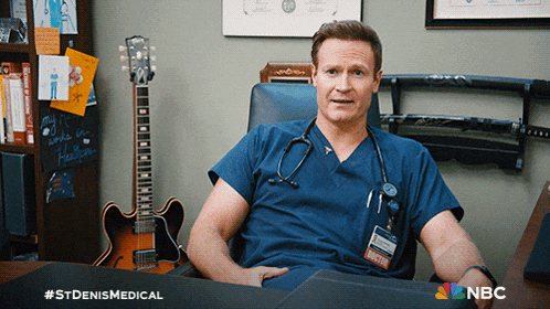 a man in a scrub sits at a desk in front of a guitar and a samurai