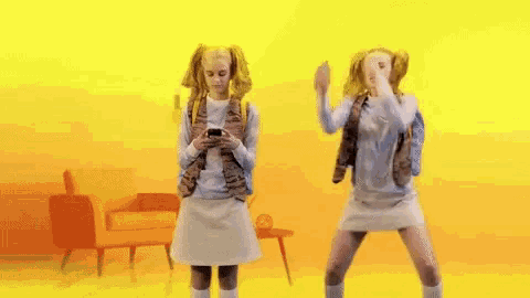 two girls are dancing in front of a yellow background while one is looking at her phone .