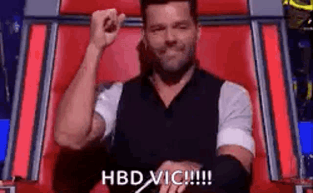 a man is sitting in a chair with his fist in the air and says `` hbd vic !!! '' .