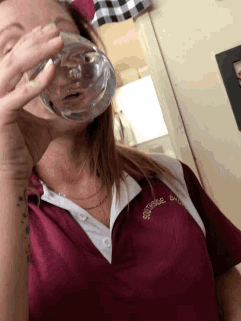 a woman wearing a maroon shirt with the word squire on it drinks from a glass