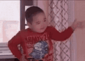 a young boy in a red shirt is standing in front of a window and making a funny face .