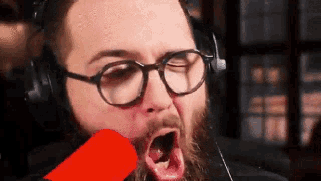 a man with glasses and a beard is wearing headphones and screaming into a microphone .