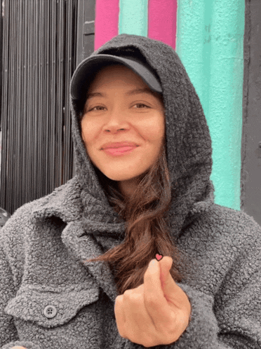 a woman wearing a hooded jacket and a hat is making a heart with her hand