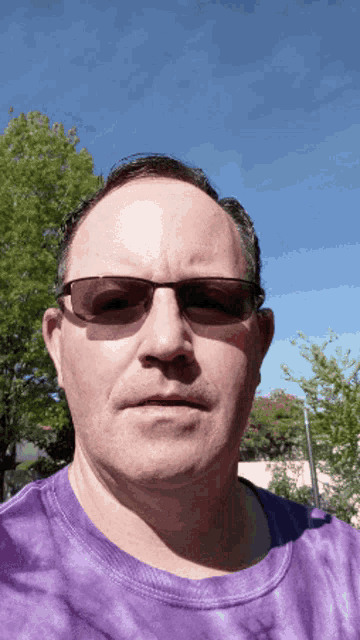 a man wearing sunglasses and a purple tie dye shirt takes a selfie