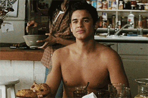 a shirtless man sits at a table with a bowl of food in front of him