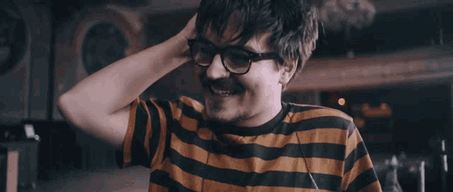 a man wearing glasses and a striped shirt smiles with his hand in his hair