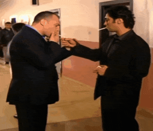 a man in a suit smoking a cigarette next to another man in a black suit