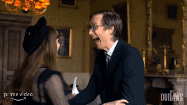 a man in a suit and tie laughs with a woman in a black dress in front of a sign that says the outlaws