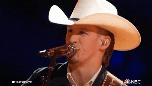 a man in a cowboy hat singing into a microphone with the nbc logo behind him