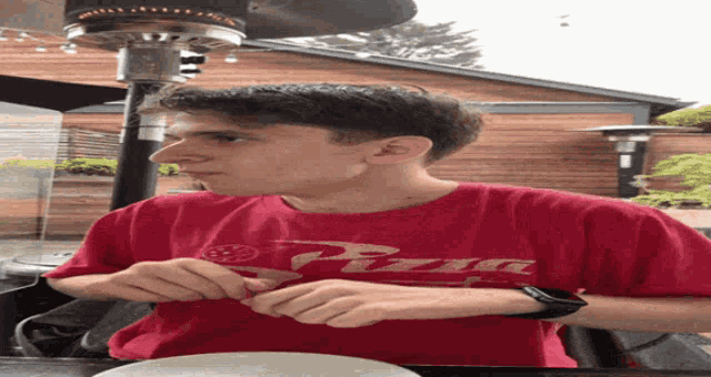 a young man wearing a red pizza shirt is sitting at a table