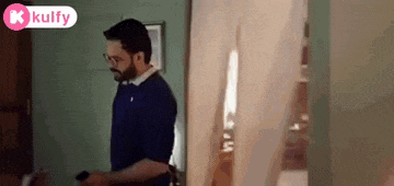 a man in a blue shirt and glasses is standing in a room holding a cell phone .