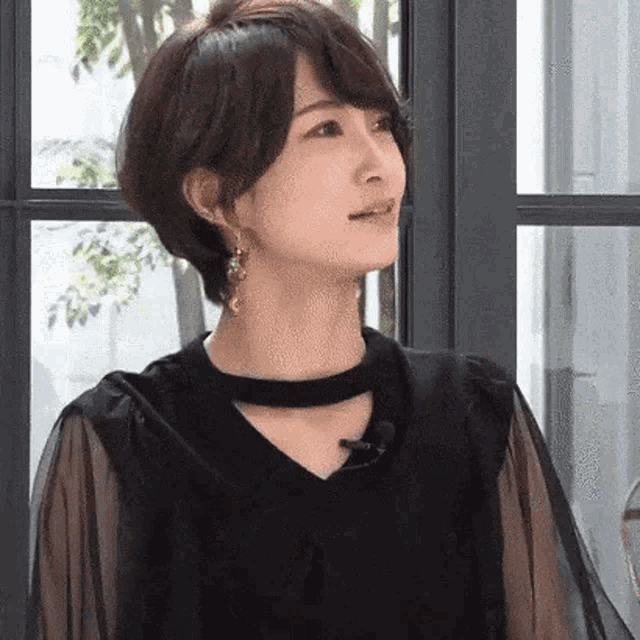 a woman wearing a black dress and earrings is standing in front of a window and looking up .