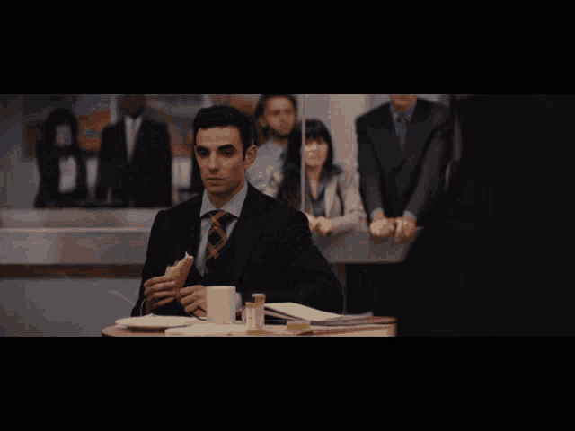 a man in a suit and tie sits at a table eating a sandwich and drinking coffee