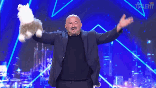 a bald man in a suit holds a stuffed animal in his hands on a stage with the word talent on the bottom