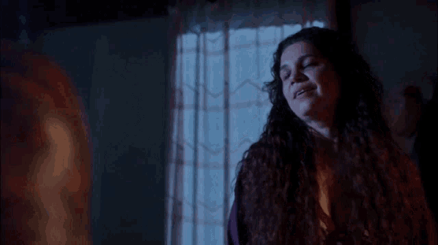 a woman with long curly hair is standing in front of a window and talking to another woman .