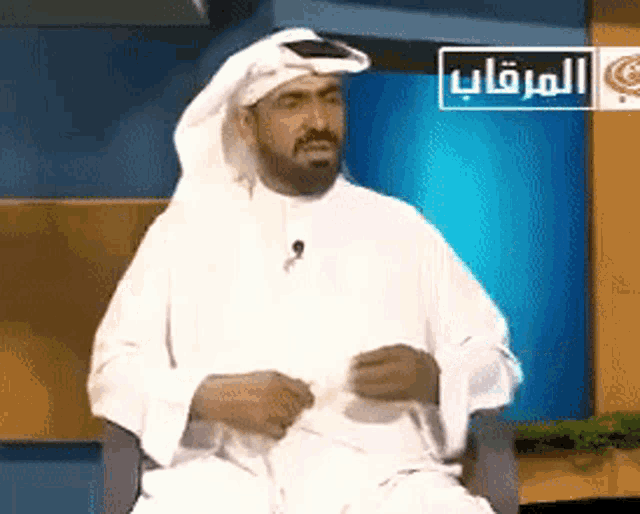 a man in a white robe is sitting in front of a blue wall with arabic writing