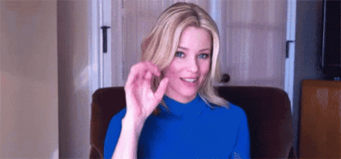 a woman in a blue sweater is sitting in a chair making a peace sign .