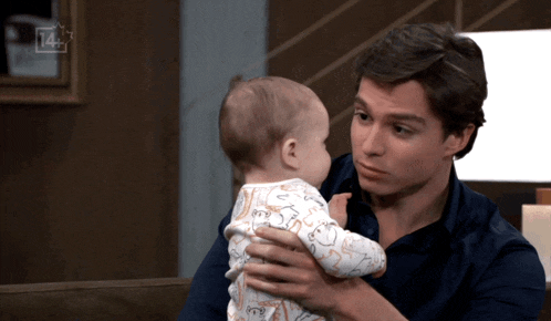 a man holds a baby in his arms with a 14+ sign in the background