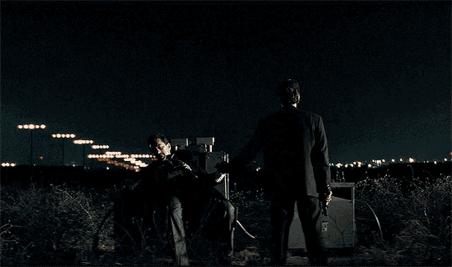 a man in a suit stands next to a man in a wheelchair in the dark