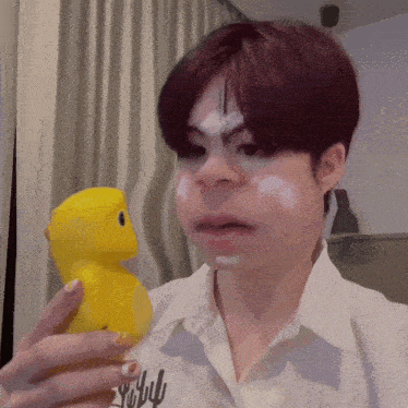 a person holding a yellow rubber duck with white paint on their face and a shirt that says " why " on it