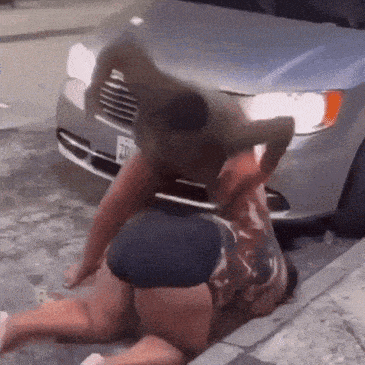 two men are fighting on the ground in front of a car