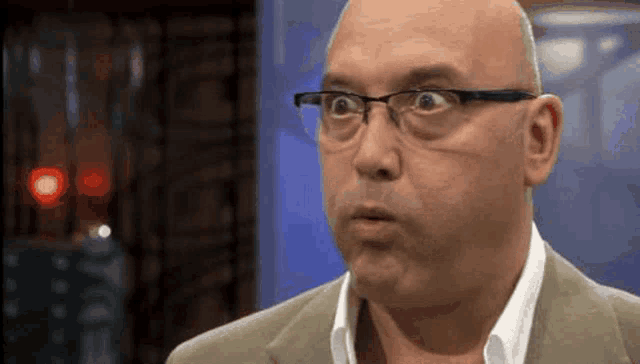 a bald man wearing glasses and a suit makes a funny face