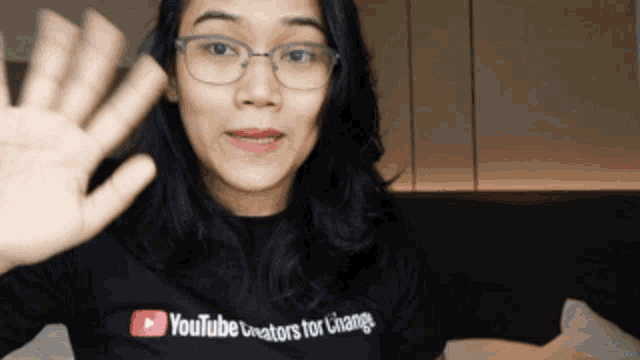 a woman wearing glasses and a youtube shirt waves her hand