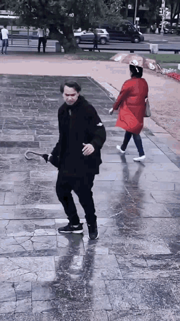 a man in a black jacket is holding a cane while a woman in a red coat walks behind him