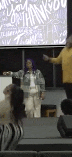 a woman with purple hair is standing in front of a group of people in front of a thank you sign .