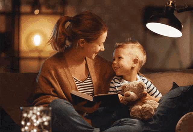 a woman sits on a couch reading a book to a young boy who is holding a teddy bear