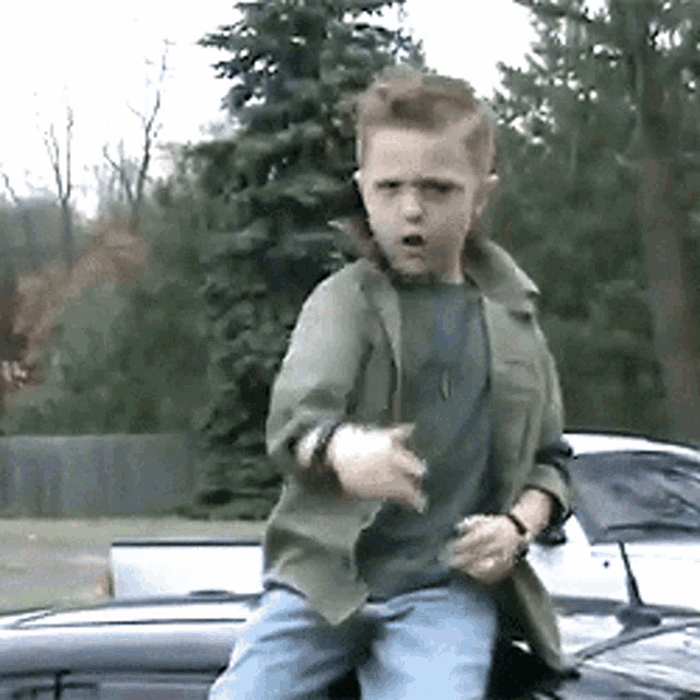 a young boy in a green jacket and blue jeans is standing on the hood of a car