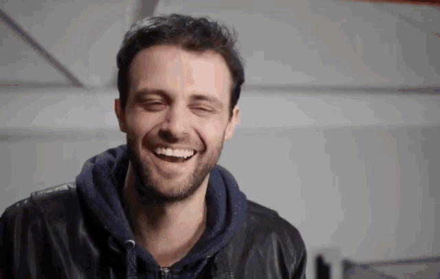 a man with a beard is smiling with his eyes closed and wearing a leather jacket .