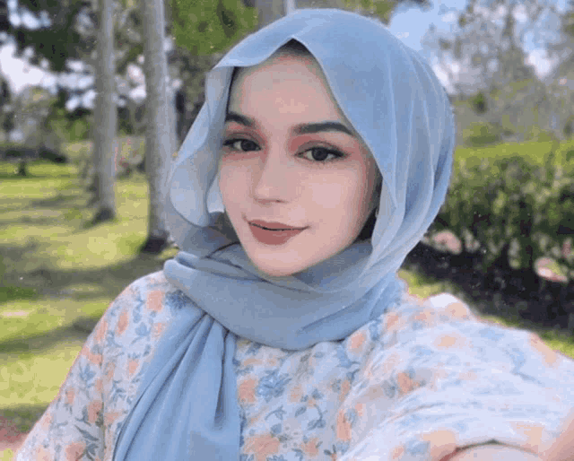 a woman wearing a light blue hijab takes a selfie in a park