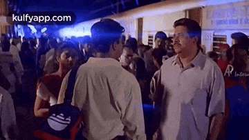 two men are standing next to each other at a train station talking to each other .