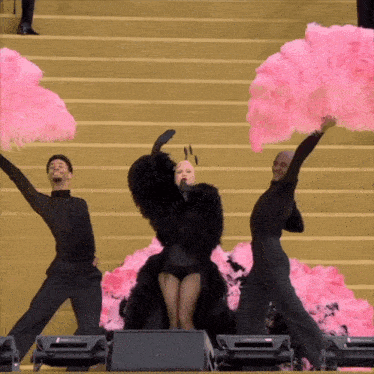 a woman in a black dress stands on a stage surrounded by pink feather fans