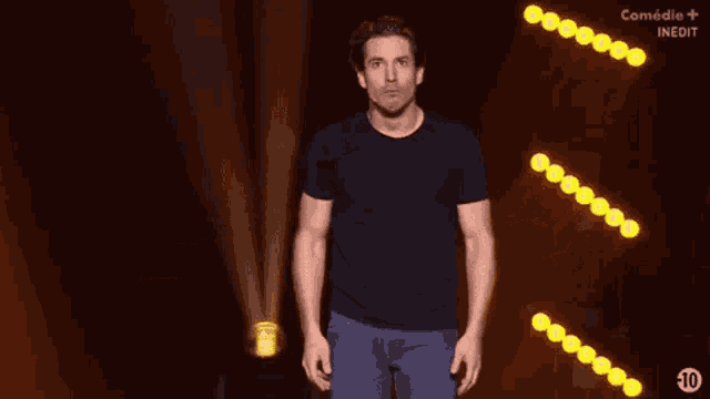 a man in a black t-shirt is standing on a stage in front of a row of yellow lights ..