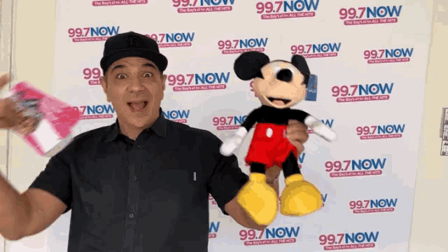 a man is holding a stuffed mickey mouse in front of a wall with 99.7 now written on it
