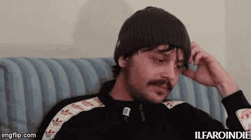 a man with a mustache is sitting on a couch wearing a beanie and a jacket .