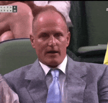 a man in a suit and tie is sitting in a stadium with a scoreboard behind him that shows the score of 674 30