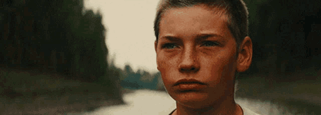 a young boy with freckles is standing in front of a river looking at the camera .