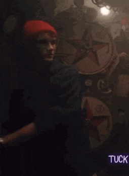 a man wearing a red hat and a watch is standing in a dark room with the word tuck on the bottom