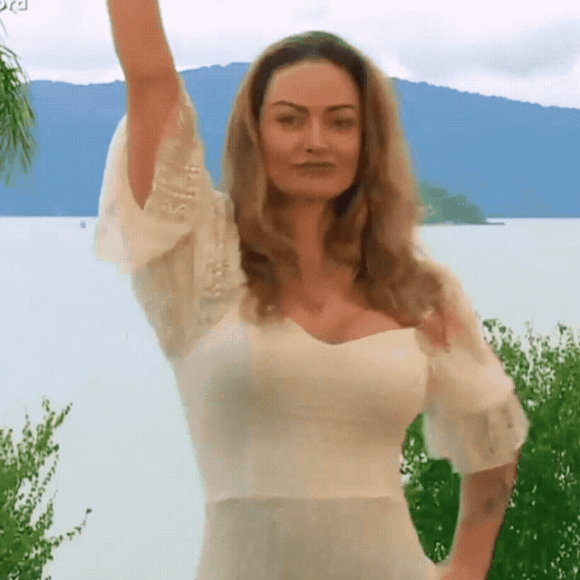 a woman in a white dress stands in front of a body of water with her arm in the air