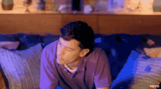 a man in a purple shirt is sitting on a blue couch with the word vevo on the bottom