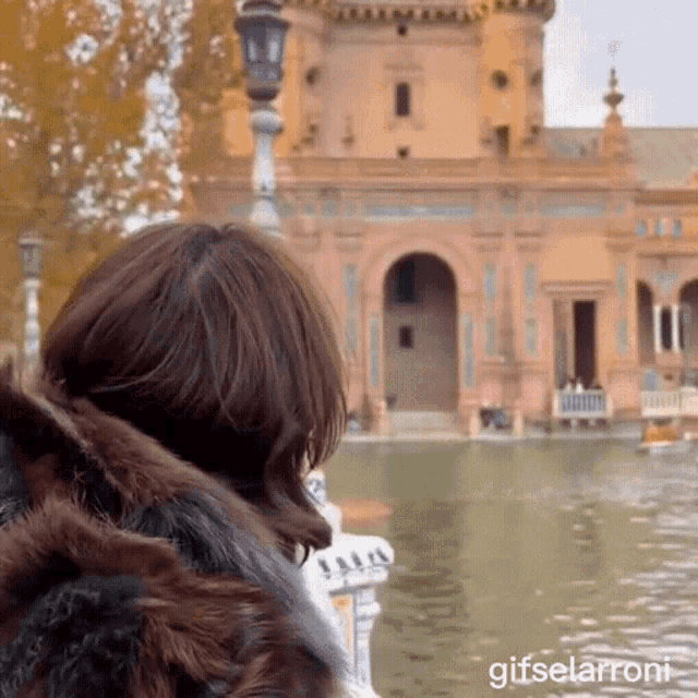 a woman in a fur coat stands in front of a building and a body of water