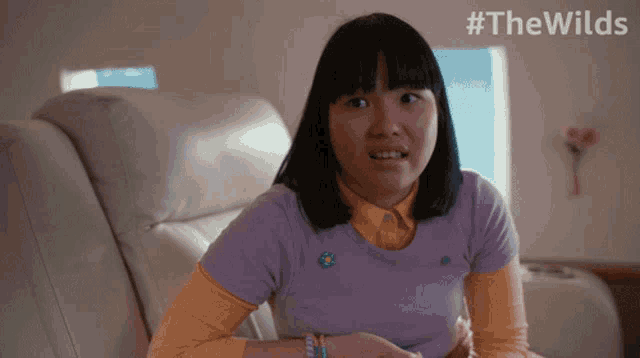 a girl in a purple shirt is sitting on a couch with the words #thewilds behind her